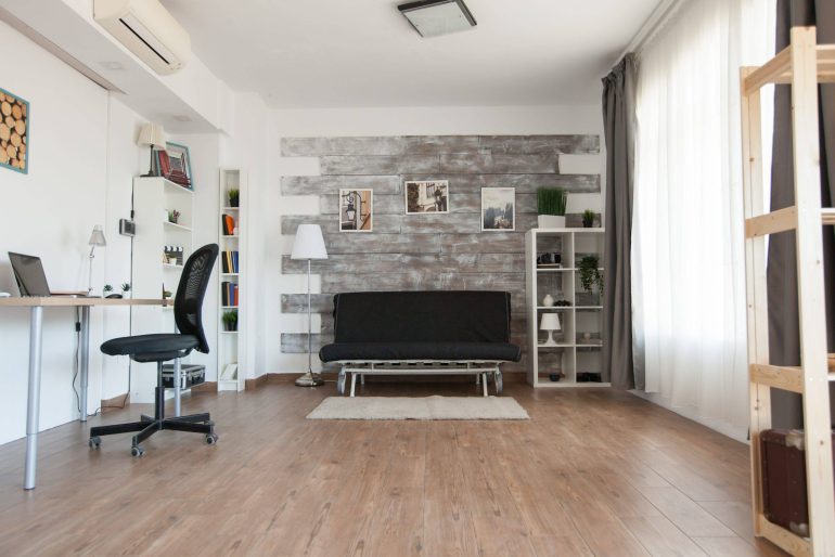 Bright home office/living area with a white desk, black swivel chair, and shelves along the left wall.